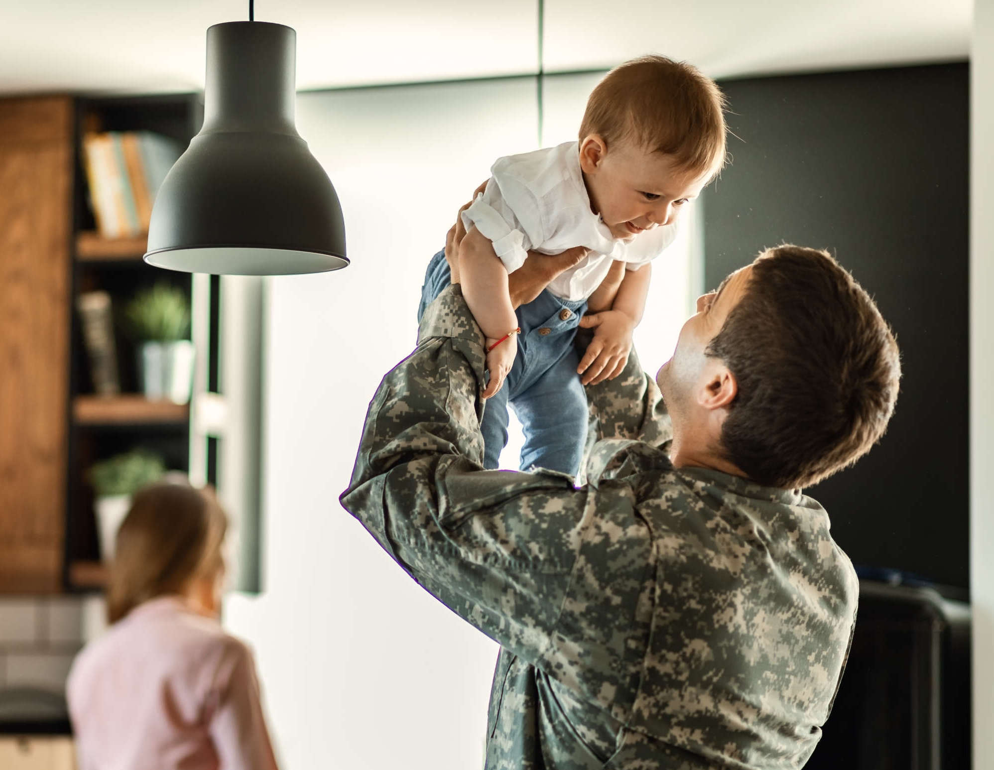 Playful military man having fun with his small son at home. Happy baby boy having fun with his military dad at home. Mother is in the background.