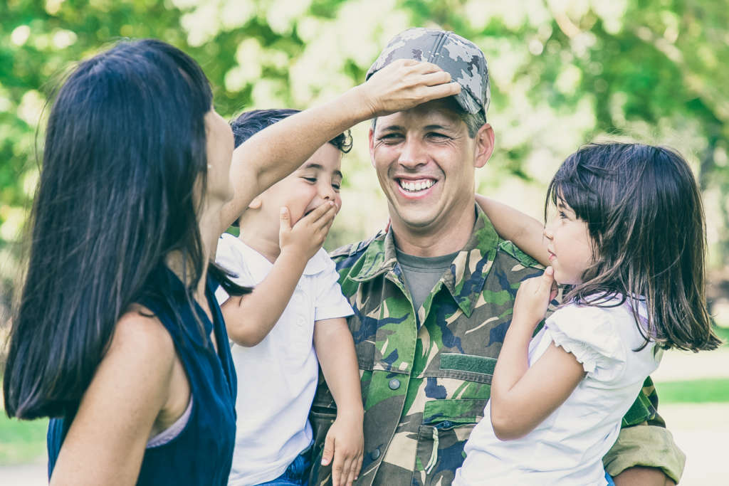 Military father with family Joyful military father in uniform returning to family, holding two kids in arms. Woman adjusting husbands cap. Family reunion or returning home concept